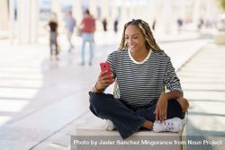 Woman smiling and checking mobile phone while sitting in pedestrian area outside 0vlkG4