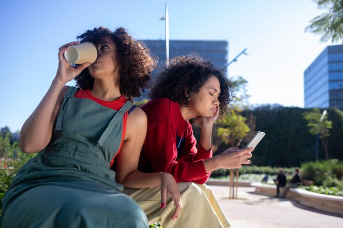 Female friends hanging in park together drinking coffee, checking phone