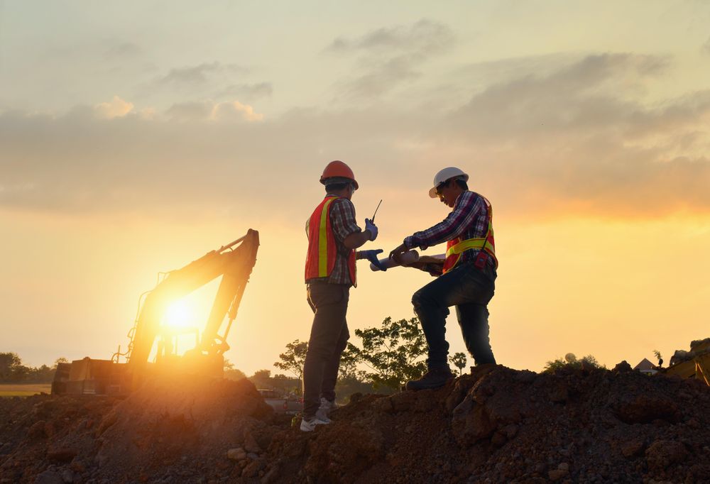Two men wearing protective helmets standing on construction site during sunset