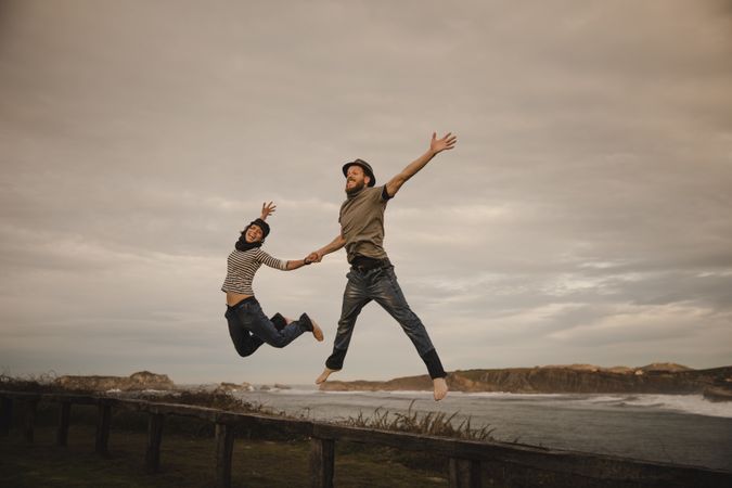 Couple holding hands and jumping on bench on shore near water
