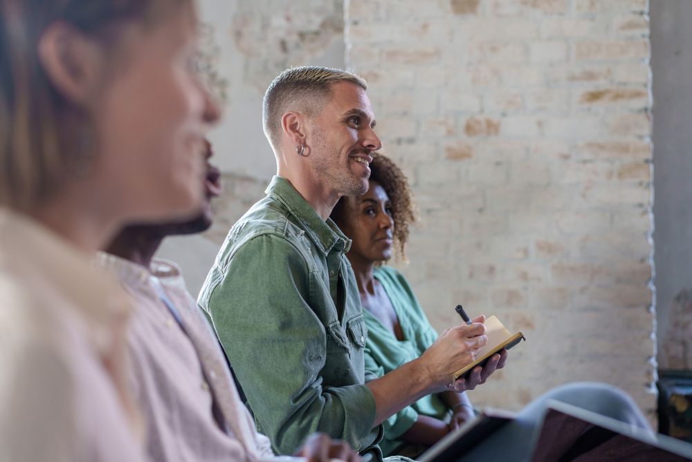 A group of people sit in rows of chairs, facing an unseen speaker, smiling and taking notes