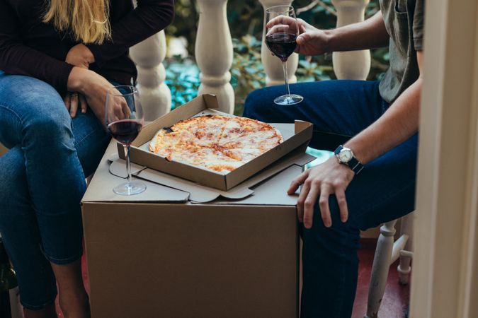 Couple sitting at home drinking wine with pizza placed on a packing box