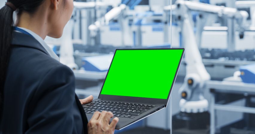 Asian Female Engineer Using Laptop Computer with Green Screen Mock Up Display. Young Woman Supervising Production at a Factory Facility with Modern Electronic Machines. Over the Shoulder Shot