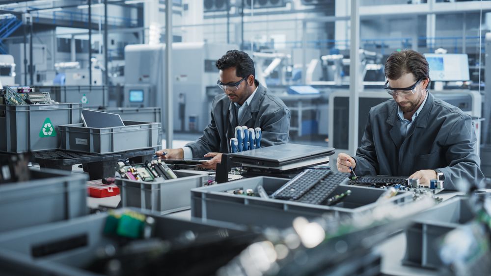 Indian And White Male Workers Taking Apart Laptops To Recycle For Production Of High Tech Devices At an Automated Electronics Factory With Robotic Arms.