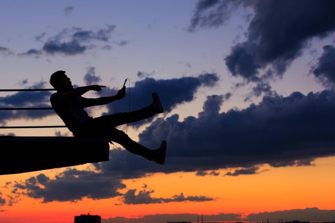 Out line of male sitting on ledge looking at tablet against a colorful dusk side, vertical