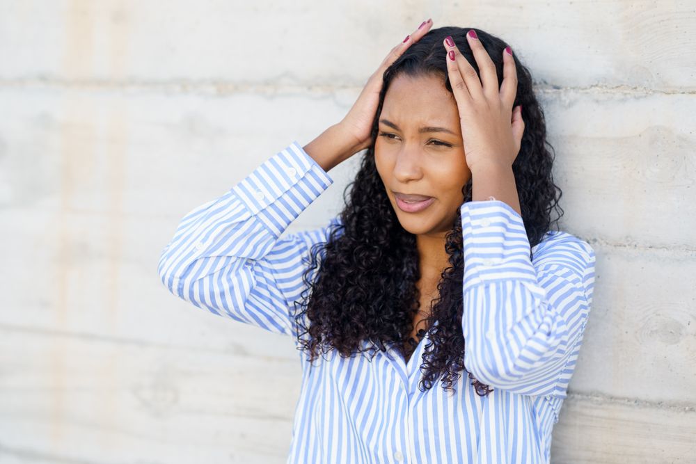 A Stressed Young Woman is Holding Her Head in Discomfort Against a Wall, Struggling With Tension