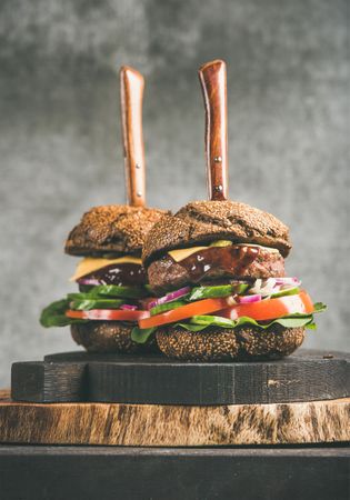 Two cheeseburgers skewered with knives, with fresh vegetables, on wooden board, vertical composition