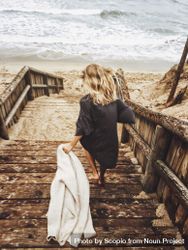 Back view of blonde woman holding a coat getting down wooden steps by seashore 4BYWX5