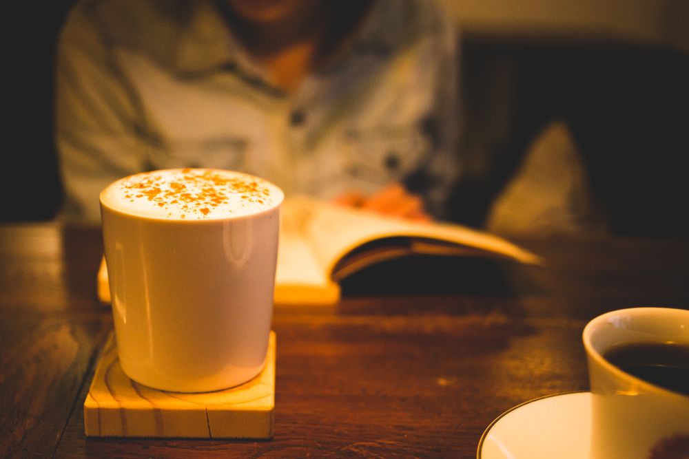 Person sitting at a table with open book and in focus mug of coffee with foam