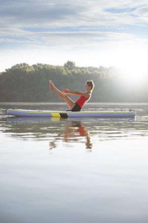 Side view of woman stretching legs and abs on paddleboard on fresh lake in the morning