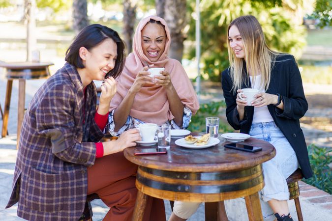 Three women laughing together while enjoying coffee and baked goods on a beautiful sunny day
