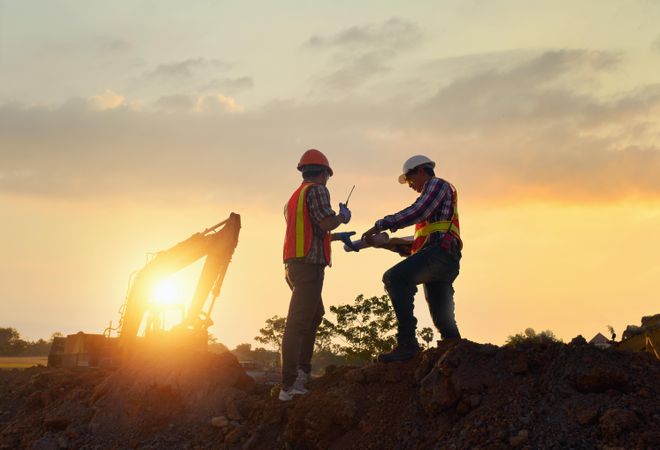 Two men wearing protective helmets standing on construction site during sunset