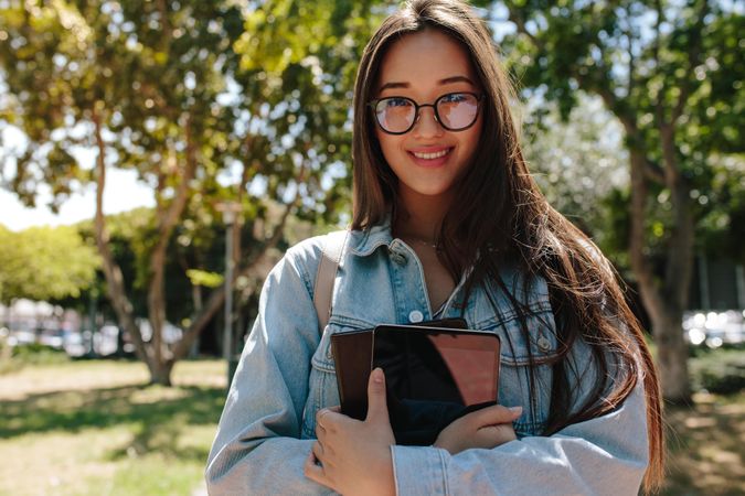 Close up of a smiling college woman standing outdoors holding tablet and book