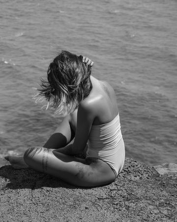 Back view of blonde woman in one piece swimsuit sitting by shoreline in grayscale