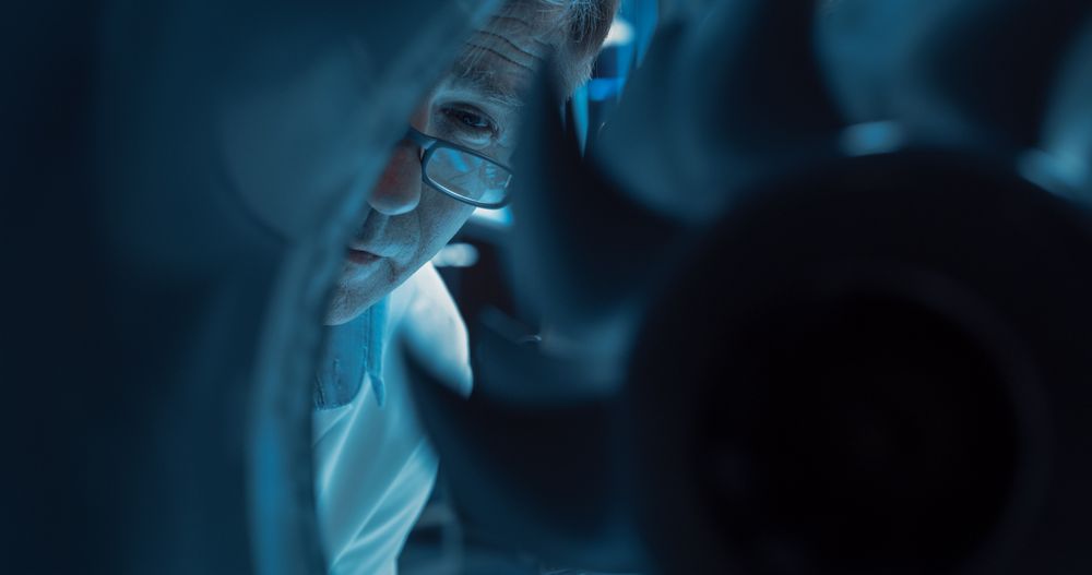 Close Up Portrait of a Mature Engineer in Glasses Examining a Rotating Fan on a High Tech Jet Engine.