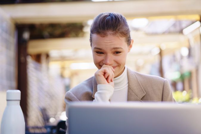 A young woman is smiling at her laptop in a modern outdoor setting, enjoying her work