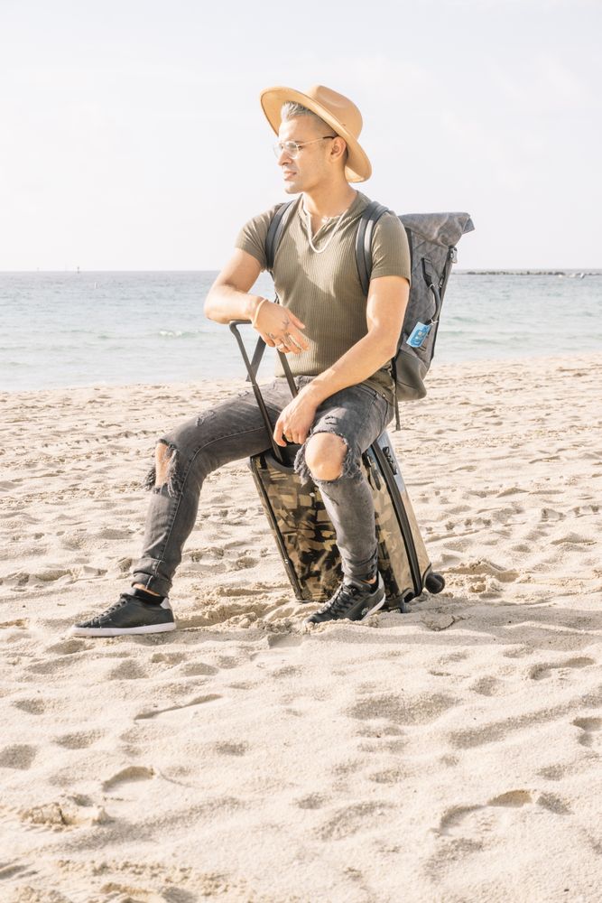 young latino man with a modern look and hat on miami beach with a travel suitcase