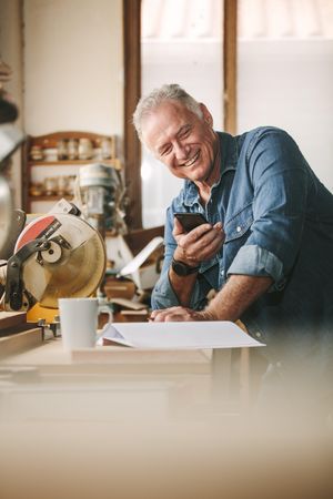 Older carpenter at his workshop using smart phone and smiling