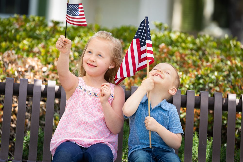 Young Sister and Brother Comparing Each Others American Flag Size On The Bench At The Park