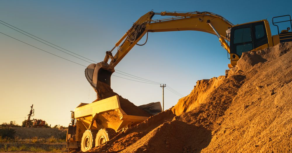 Construction Site On Sunny Evening: Industrial Excavator Loading Sand Into A Truck. The Process Of Building New Apartment Complex. Workers Operating Heavy Machinery To Complete New Real Estate Project