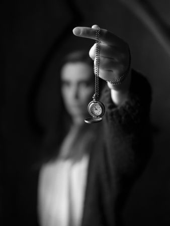 A young woman, out of focus, holds a pocket watch on a chain, and shows it hanging in the air, while time passes