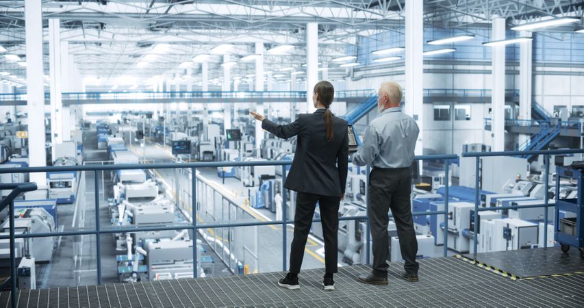 Male Engineer and Female Project Manager Standing on a Platform, Talking, Using Laptop Computer and Monitoring Production at a Modern Automated Electronics Manufacture with AI Robotic Arms