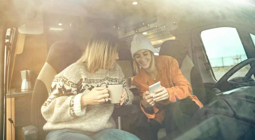 Female friends sitting in parked van having coffee and checking social media on phone