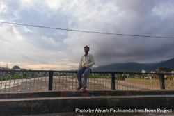 Indian man sitting on railing of bridge over shallow river on cloudy day in Dehradun, India bE9mVb