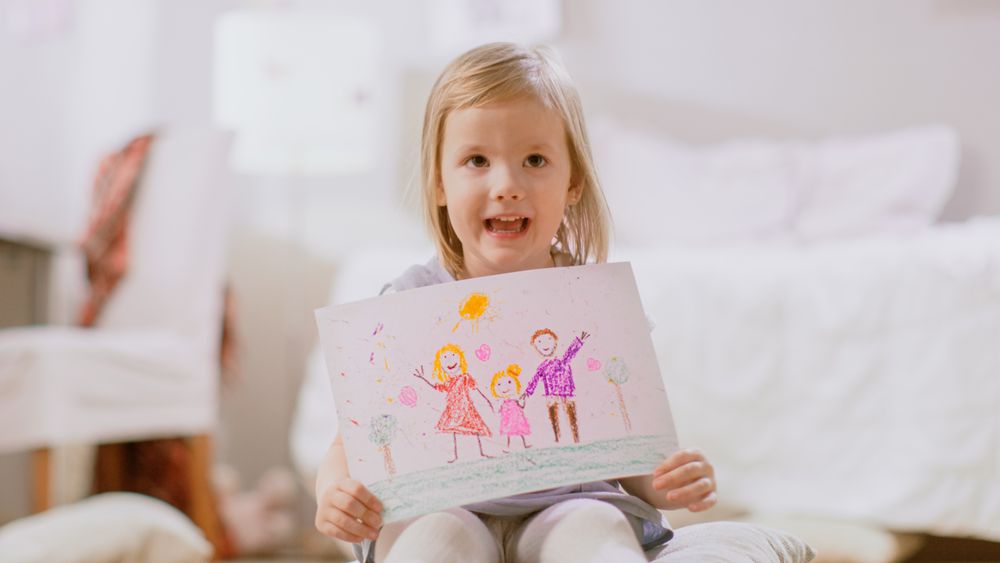 Young Girl Sitting on Pillows and Shows Drawing of Her Family.