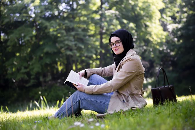 Smiling woman in headscarf sitting on the grass with a book