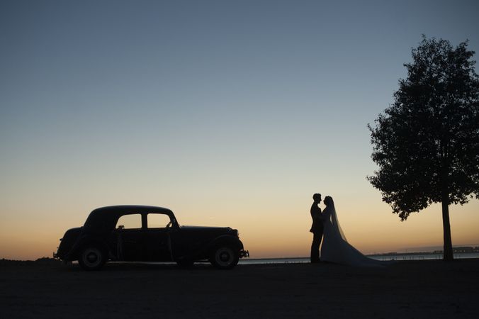 Woman and man holding hands near vintage car at sunset