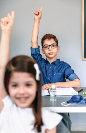 Students with mask on table raising hands at school