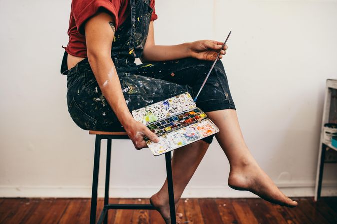 Copped side view of woman sitting on a stool holding painting tools in studio