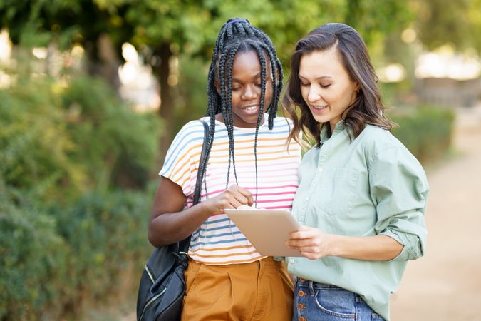 Women friends pointing at something on tablet in casual clothing in park