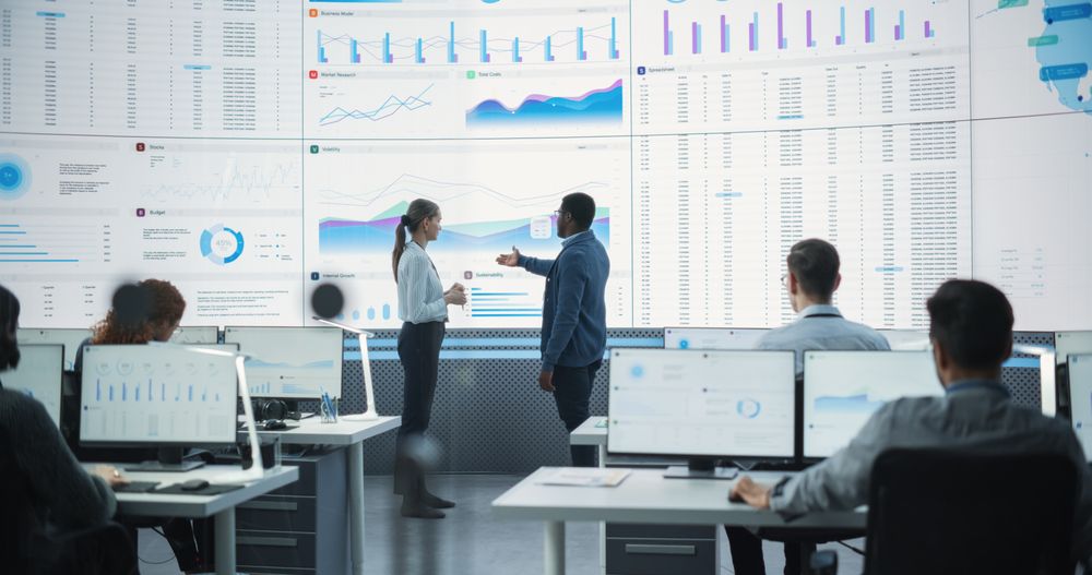 Black Male Financial Data Analysts And White Female Project Manager Having A Meeting In Monitoring Office With Analytics Feed On Big Digital Screen.