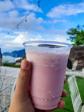 Woman's hand holding a plastic glass of strawberry milkshake on outdoor cafe background