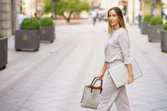 Chic woman in grey walking with laptop outside