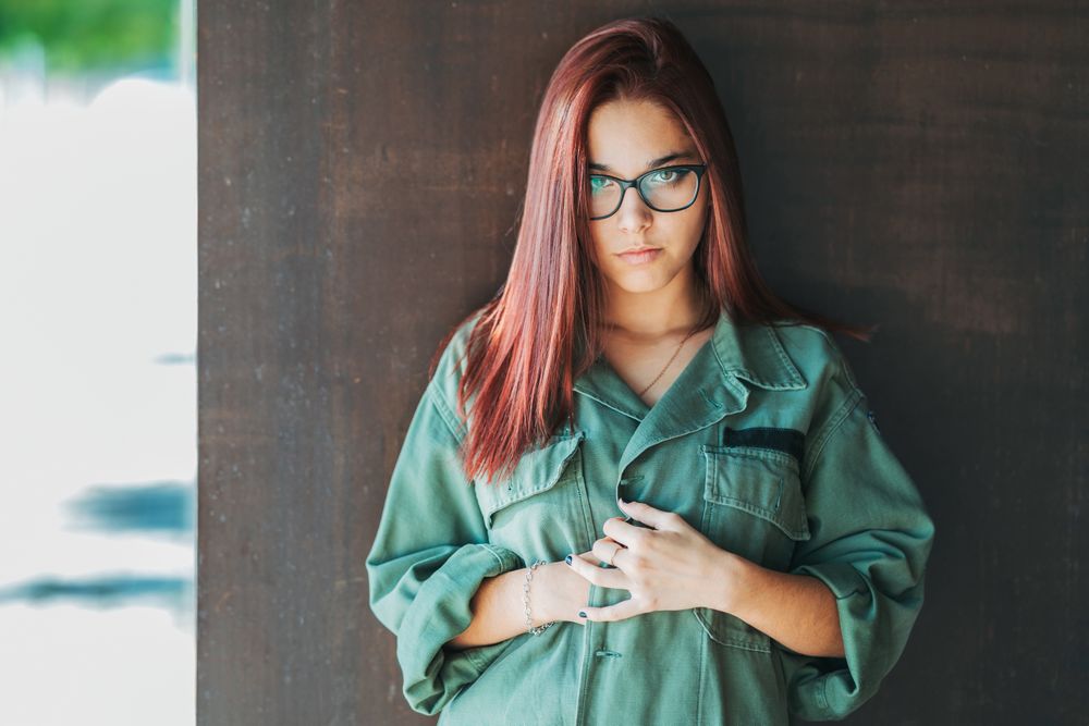 Stylish confident bright woman in glasses leaning on wall and looking at camera