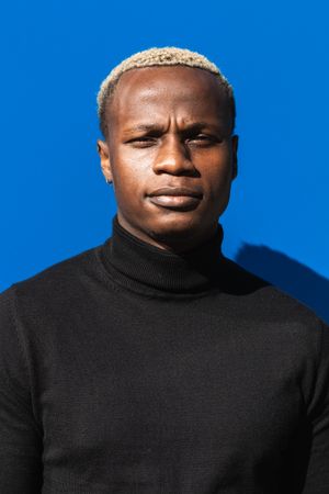 Serious Black guy standing against blue wall in sunlight