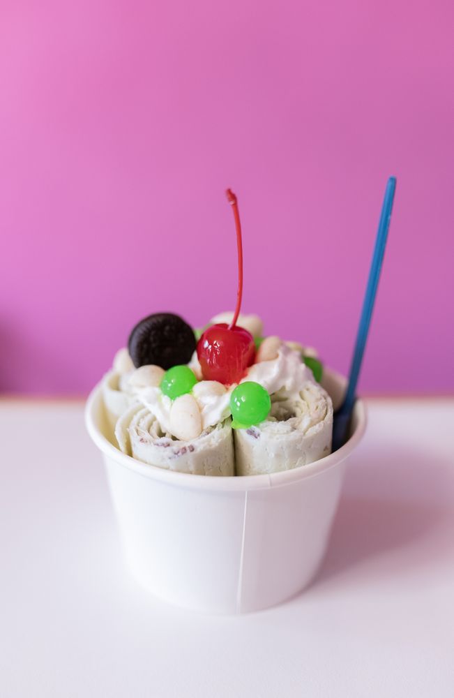 Close up of a rolled ice cream decorated with a cherry, a cookie and green bubble toppings