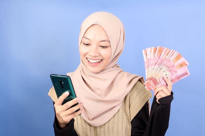 Young Asian Muslim woman in Brown Shirt using Mobile Phone and Holding Cash Money