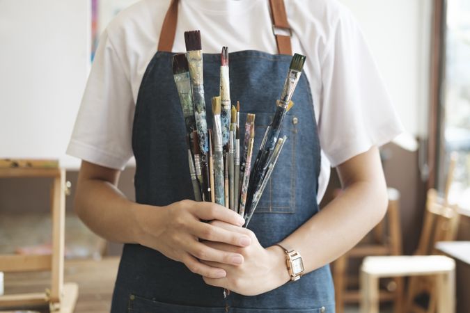 A close-up of the artist hands wearing an apron smeared with paint. Clutching many brushes and paintbrushes.