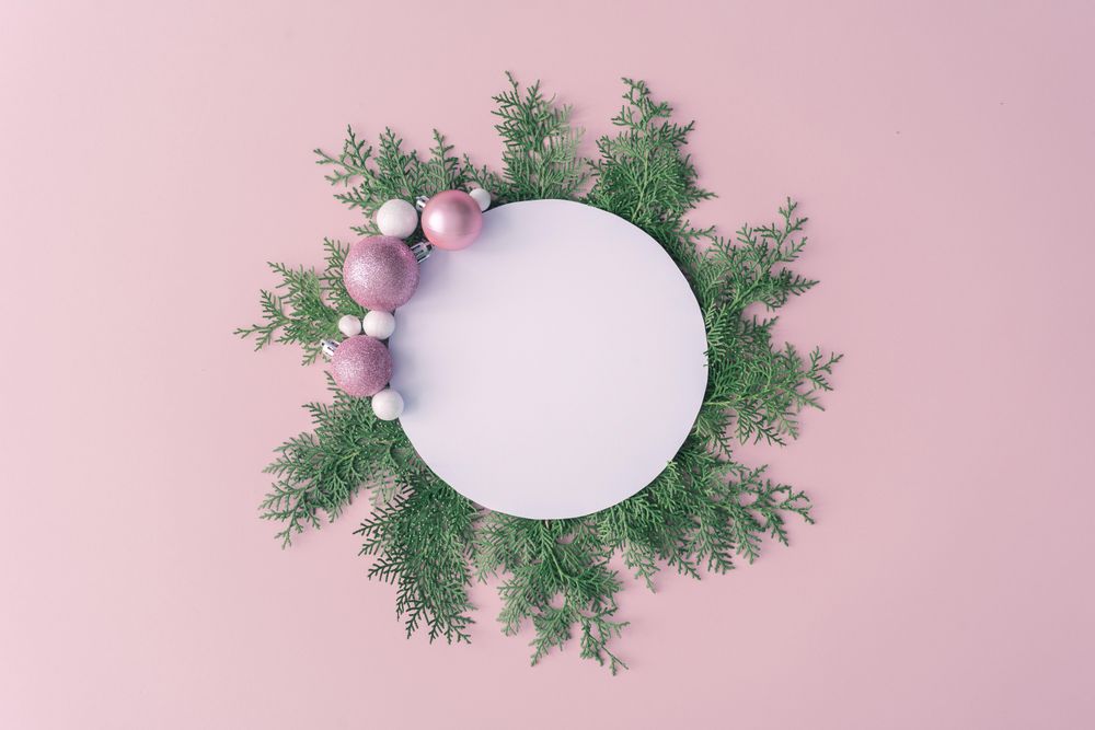 Christmas frame made of winter plants and pink baubles on pink table background