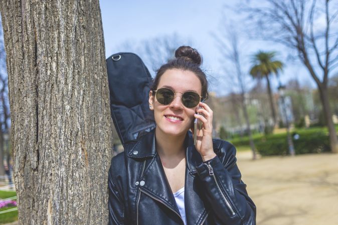 Side view of a young laughing hipster woman standing and leaning on a tree at park in sunny day while using a mobile phone