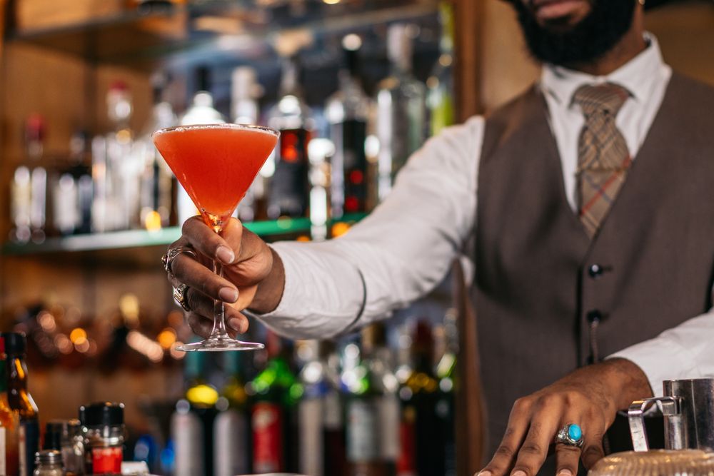 Cutout of a Black bartender offering a cocktail served in a martini glass in a traditional cocktail bar
