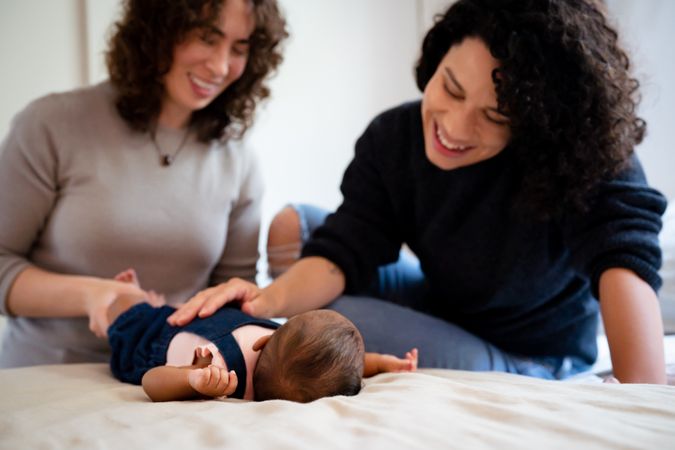 Two smiling new moms looking down at infant on bed with hand on baby’s tummy