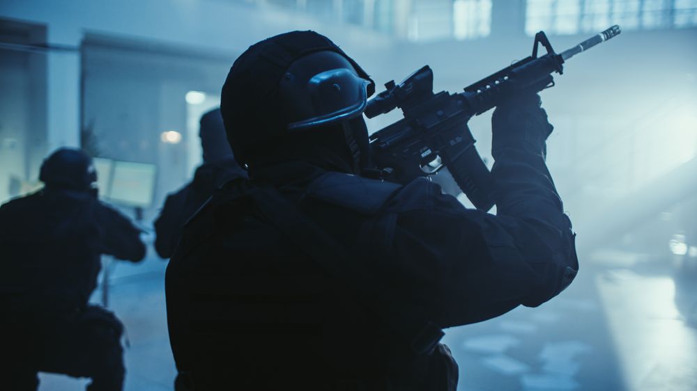 Portrait of Masked Armed SWAT Police Officer Storming Dark Seized Office Building with Desks and Computers.