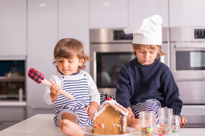 Two kids sitting on kitchen counter making gingerbread house