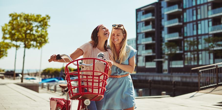 Two close female friends happily walking along a riverwalk with red bike on sunny day