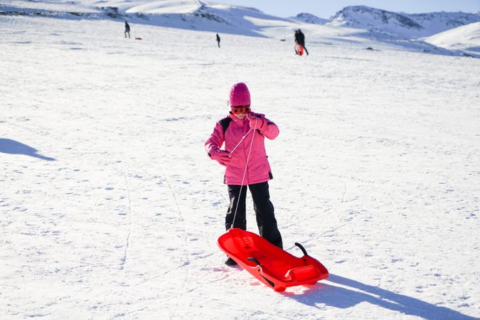 Child in pink snow suit standing with sled at resort